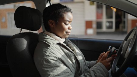 Téléchargez les photos : Femme afro-américaine en utilisant smartphone assis sur la voiture à la rue - en image libre de droit