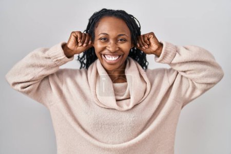 Téléchargez les photos : Femme africaine debout sur fond blanc souriant tirant les oreilles avec les doigts, geste drôle. problème d'audition - en image libre de droit