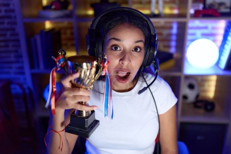 Foto de Mujer árabe joven jugando videojuegos sosteniendo trofeo asustado y sorprendido con la boca abierta para sorpresa, cara de incredulidad - Imagen libre de derechos