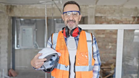 Téléchargez les photos : Moyen âge homme constructeur souriant confiant tenant hardhat sur le chantier de construction - en image libre de droit