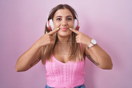 Téléchargez les photos : Jeune femme blonde écoutant de la musique à l'aide d'écouteurs souriant avec la bouche ouverte, les doigts pointant et forçant sourire joyeux - en image libre de droit