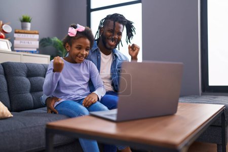 Téléchargez les photos : Père et fille africains faisant appel vidéo sur ordinateur portable à la maison criant fiers, célébrant la victoire et le succès très excité avec le bras levé - en image libre de droit