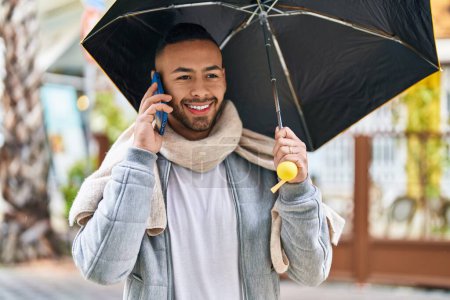 Foto de African american man talking on smartphone holding umbrella at street - Imagen libre de derechos