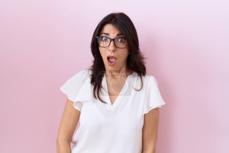 Téléchargez les photos : Femme hispanique du Moyen-âge portant un t-shirt blanc décontracté et des lunettes au visage choqué, à l'air sceptique et sarcastique, surprise avec la bouche ouverte - en image libre de droit