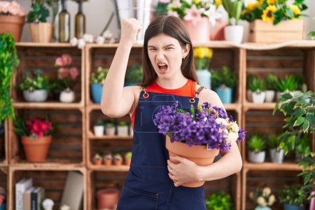 Foto de Joven mujer caucásica que trabaja en floristería sosteniendo maceta con flores molestas y frustradas gritando de rabia, gritando loca de rabia y levantando la mano - Imagen libre de derechos