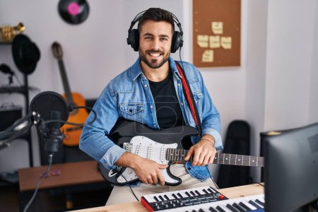 Téléchargez les photos : Jeune homme hispanique souriant confiant tenant la guitare électrique au studio de musique - en image libre de droit