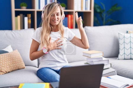 Téléchargez les photos : Jeune femme blonde étudiant à l'aide d'un ordinateur portable à la maison souriant jurer avec la main sur la poitrine et les doigts vers le haut, faire un serment de fidélité - en image libre de droit