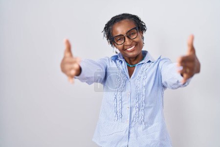 Téléchargez les photos : Femme africaine avec dreadlocks debout sur fond blanc portant des lunettes regardant la caméra souriant à bras ouverts pour étreindre. expression joyeuse embrassant le bonheur. - en image libre de droit