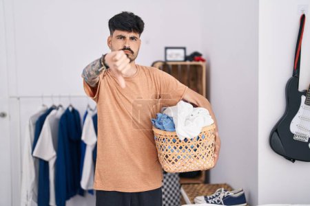 Photo for Young hispanic man with beard holding laundry basket at bedroom with angry face, negative sign showing dislike with thumbs down, rejection concept - Royalty Free Image