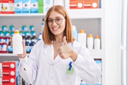 Téléchargez les photos : Jeune rousse femme travaillant à la pharmacie pharmacie tenant écran solaire souriant heureux et positif, pouce levé faire excellent et signe d'approbation - en image libre de droit