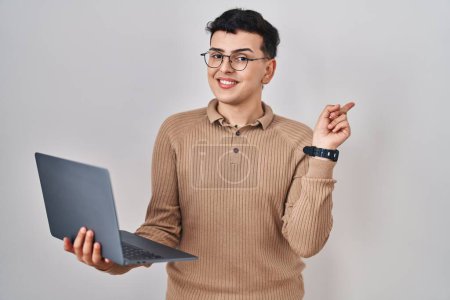 Photo for Non binary person using computer laptop with a big smile on face, pointing with hand finger to the side looking at the camera. - Royalty Free Image
