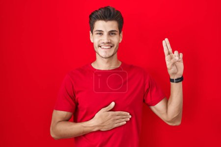 Téléchargez les photos : Jeune homme hispanique debout sur fond rouge souriant jurer avec la main sur la poitrine et les doigts vers le haut, faisant un serment de fidélité promesse - en image libre de droit