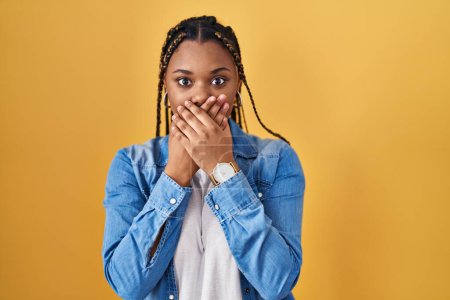 Photo for African american woman with braids standing over yellow background shocked covering mouth with hands for mistake. secret concept. - Royalty Free Image