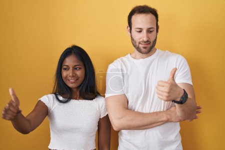 Téléchargez les photos : Couple interracial debout sur fond jaune regardant fier, souriant faire les pouces vers le haut geste sur le côté - en image libre de droit