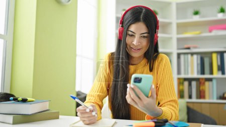 Téléchargez les photos : Jeune belle étudiante hispanique écrivant des notes à l'aide d'un smartphone et d'écouteurs à la bibliothèque universitaire - en image libre de droit