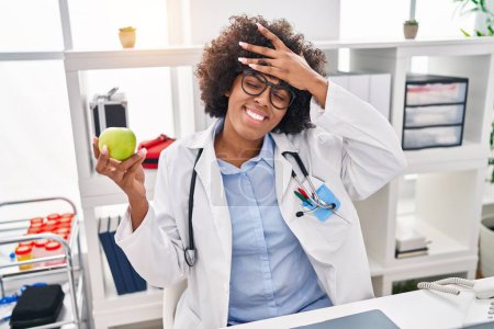 Photo for Black doctor woman with curly hair holding green apple stressed and frustrated with hand on head, surprised and angry face - Royalty Free Image
