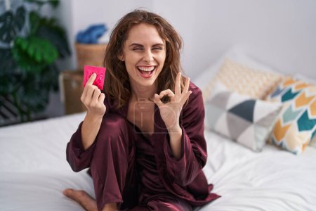 Téléchargez les photos : Belle femme brune tenant un préservatif dans la chambre à coucher faisant ok signe avec les doigts, souriant geste amical excellent symbole - en image libre de droit