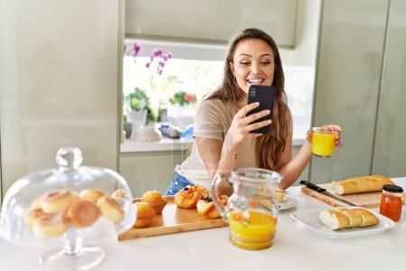 Foto de Joven hermosa mujer hispana desayunando usando smartphone en la cocina - Imagen libre de derechos