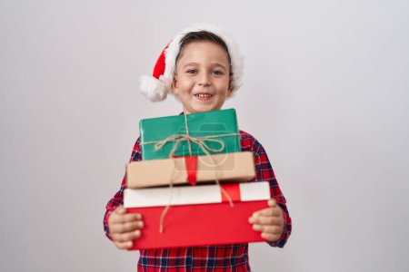 Foto de Pequeño niño hispano con sombrero de navidad sosteniendo regalos sonriendo y riendo en voz alta porque divertido chiste loco. - Imagen libre de derechos