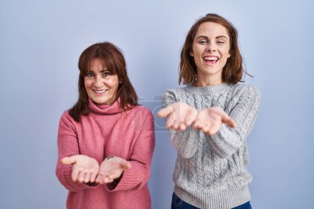 Téléchargez les photos : Mère et fille debout sur fond bleu souriant avec les mains paumes ensemble recevant ou donnant geste. maintien et protection - en image libre de droit