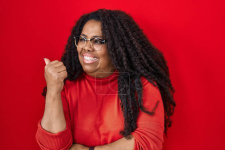 Photo for Plus size hispanic woman standing over red background smiling with happy face looking and pointing to the side with thumb up. - Royalty Free Image