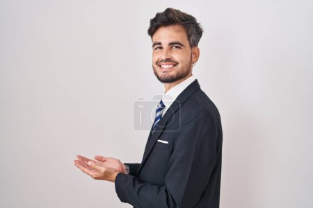 Foto de Joven hombre hispano con tatuajes vistiendo traje de negocios y corbata apuntando a un lado con las manos abiertas mostrando espacio de copia, presentando anuncio sonriente emocionado feliz - Imagen libre de derechos