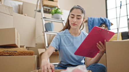 Photo for Young beautiful hispanic woman unpacking cardboard box writing on checklist at new home - Royalty Free Image