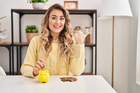 Photo for Young caucasian woman sitting on the table with piggy bank and coins pointing thumb up to the side smiling happy with open mouth - Royalty Free Image