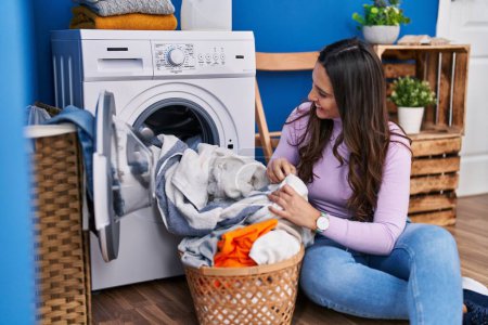 Photo for Young hispanic woman washing clothes sitting on floor at laundry room - Royalty Free Image