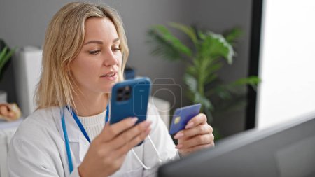 Foto de Mujer rubia joven médico de compras con teléfono inteligente y tarjeta de crédito en la clínica - Imagen libre de derechos