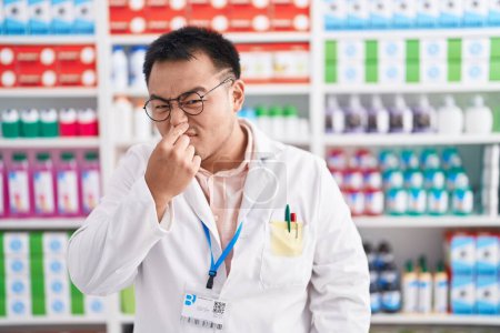 Foto de Joven chino trabajando en farmacia oliendo algo apestoso y asqueroso, olor intolerable, conteniendo la respiración con los dedos en la nariz. mal olor - Imagen libre de derechos