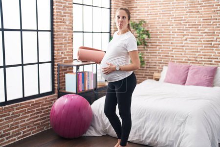 Photo for Young pregnant woman holding pilate mat at bedroom making fish face with mouth and squinting eyes, crazy and comical. - Royalty Free Image