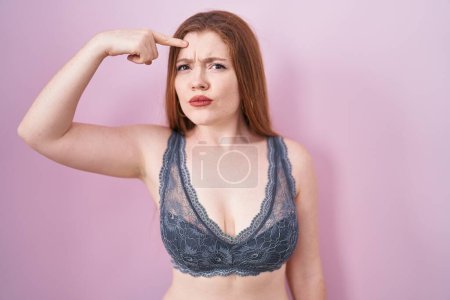 Photo for Redhead woman wearing lingerie over pink background pointing unhappy to pimple on forehead, ugly infection of blackhead. acne and skin problem - Royalty Free Image