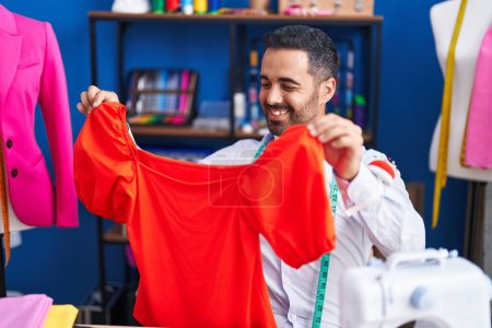 Foto de Joven hombre hispano sastre sonriendo confiado sosteniendo la ropa en el estudio de costura - Imagen libre de derechos