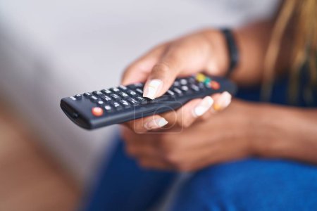 African american woman holding tv remote control sitting on sofa at home