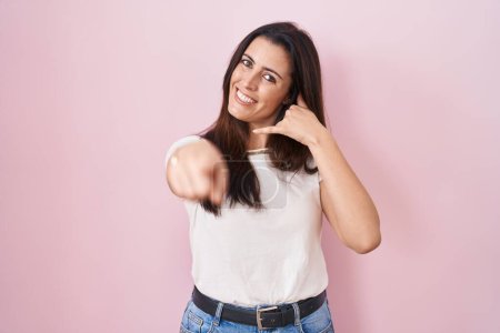 Photo for Young brunette woman standing over pink background smiling doing talking on the telephone gesture and pointing to you. call me. - Royalty Free Image