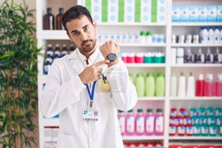 Téléchargez les photos : Bel homme hispanique travaillant à la pharmacie pharmacie à la hâte pointant vers regarder le temps, l'impatience, regardant la caméra avec une expression détendue - en image libre de droit
