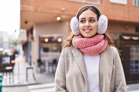 Téléchargez les photos : Jeune belle femme hispanique souriant confiant portant écharpe et cache-oreilles dans la rue - en image libre de droit