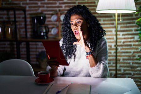 Photo for Middle age hispanic woman using touchpad sitting on the table at night hand on mouth telling secret rumor, whispering malicious talk conversation - Royalty Free Image
