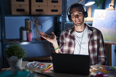 Téléchargez les photos : Jeune homme hispanique assis au studio d'art avec ordinateur portable tard dans la nuit souriant gai présentation et pointant avec la paume de la main regardant la caméra. - en image libre de droit