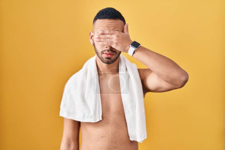 Téléchargez les photos : Jeune homme hispanique debout torse nu avec serviette couvrant les yeux avec la main, l'air sérieux et triste. concept de non-visibilité, de dissimulation et de rejet - en image libre de droit