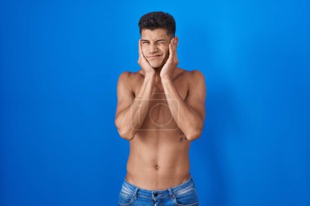Photo for Young hispanic man standing shirtless over blue background covering ears with fingers with annoyed expression for the noise of loud music. deaf concept. - Royalty Free Image