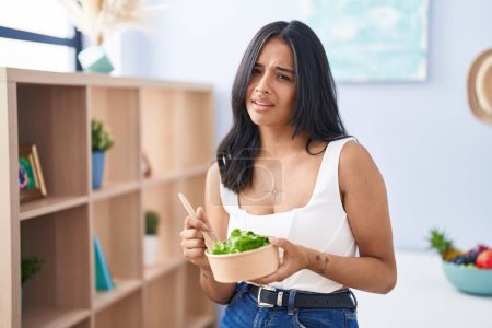 Téléchargez les photos : Femme brune mangeant une salade à la maison expression inutile et confuse. concept de doute. - en image libre de droit