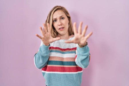 Photo for Young blonde woman standing over pink background afraid and terrified with fear expression stop gesture with hands, shouting in shock. panic concept. - Royalty Free Image