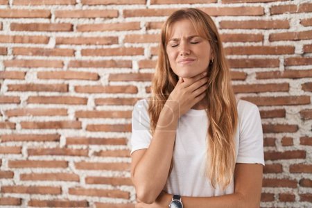 Foto de Mujer caucásica joven de pie sobre la pared de ladrillos tocando el cuello doloroso, dolor de garganta para la gripe, la clod y la infección - Imagen libre de derechos