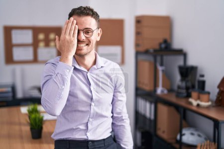 Foto de Joven hombre hispano en la oficina cubriendo un ojo con la mano, sonrisa confiada en la cara y emoción sorpresa. - Imagen libre de derechos