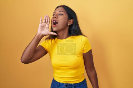 Téléchargez les photos : Jeune femme indienne debout sur fond jaune criant et criant fort à côté avec la main sur la bouche. concept de communication. - en image libre de droit