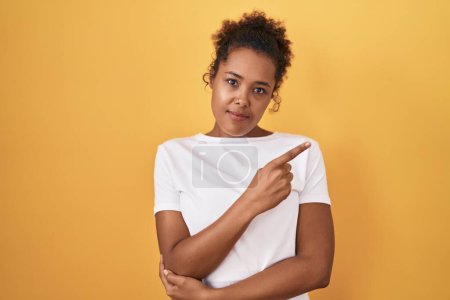 Téléchargez les photos : Jeune femme hispanique avec les cheveux bouclés debout sur fond jaune pointant avec le doigt de la main sur le côté montrant la publicité, visage sérieux et calme - en image libre de droit
