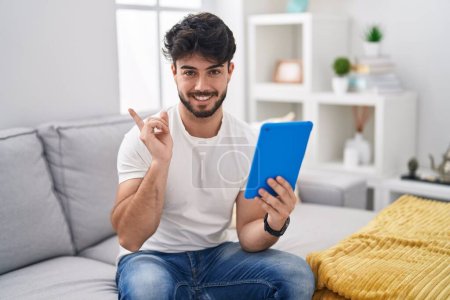 Photo for Hispanic man with beard using touchpad sitting on the sofa with a big smile on face, pointing with hand finger to the side looking at the camera. - Royalty Free Image