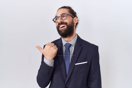 Photo for Hispanic man with beard wearing suit and tie smiling with happy face looking and pointing to the side with thumb up. - Royalty Free Image